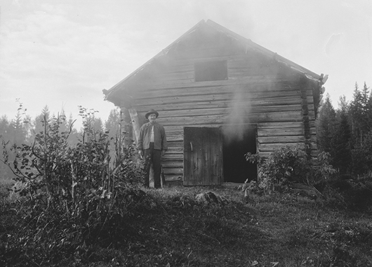 Johan Olssoni Röjdfors framför sin bastu. Bastun saknar skorsten. Röken tränger ut från dörröppningen. Foto: Astrid Reponen 1930, Museiverkets bildsamlingar SUK334 70