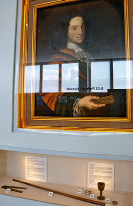 Under porträttet av Polhem syns en proportionalcirkel likadan som den han håller i handen, hans käpp och snusdosa, en bägare och en medaljong.