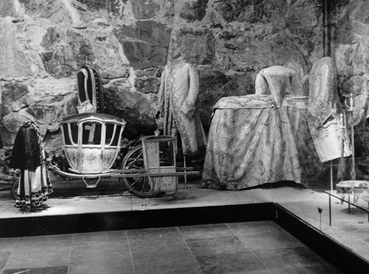 Drottningars och prinsessors kläder började sparas först vid mitten av 1700- talet, och var sällsynta i den ursprungliga basutställningen. I denna scenbild dominerar museets första klänning, drottning Sofia Magdalenas brudklänning i blommig silverbrokad broderad med knypplade silverspetsar och, för säkerhets skull, omgiven av tre av maken, Gustav III:s kostymer, längst t v serafimerkolt för den tvåårige kronprinsen, hans myndighetsdräkt och slutligen, hans i Paris sydda bröllopsdress i silverbrokad, broderad med guldtråd och blåskimrande paljetter. I Paris hade 40 personer dag och natt i nära 4 veckors tid arbetat med dressen. Det blev en engångsdräkt. Den händelserika scenbilden utspelar sig mot en vägg av murad grovhuggen gråsten och ett golv av slitstark, gotländsk kalksten.